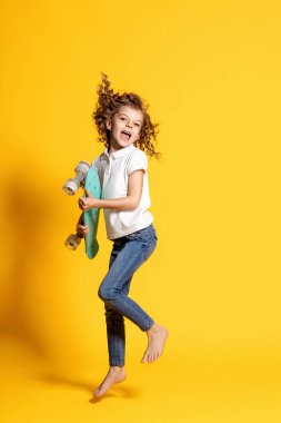Full length of curly emotional little girl wearing white polo, blue jeans, jumping with skateboard in hands while isolated on yellow background. Space for advertising 