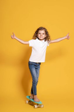 Full length of curly funny little girl wearing white polo, blue jeans, who skating on penny board and gesturing thumbs up while isolated on yellow background. Space for advertising 
