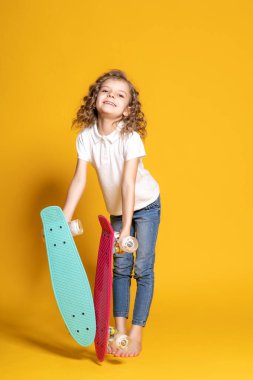 Full length of curly happy little girl in white polo, blue jeans who holding two colorful skateboards isolated on yellow background. Space for advertising 