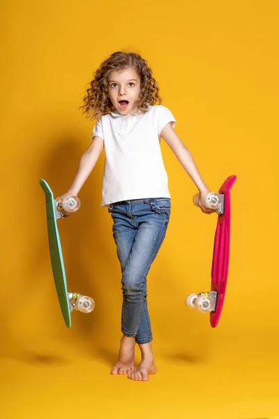 Full length of surprised little curly girl who holds two colorful skateboards and doesnt know which one to choose on yellow background. Problem of choice
