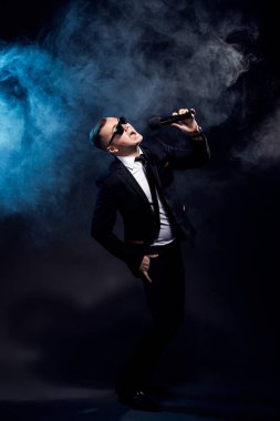 Full length of stylish young man in sunglasses and black suit singing in microphone against a dark background with smoke, actor, singer, show, host of the event. 