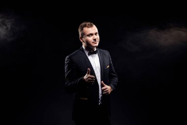Handsome confident businessman wearing suit with bow tie gesturing thumbs up isolated over black background. Space for advertising 