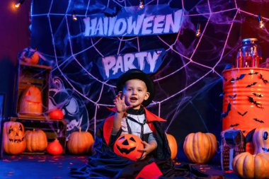 Halloween party. Little cute boy in Dracula costume at Halloween party sitting against Halloween decorations and holds pumpkin for candies. Trick or treat 