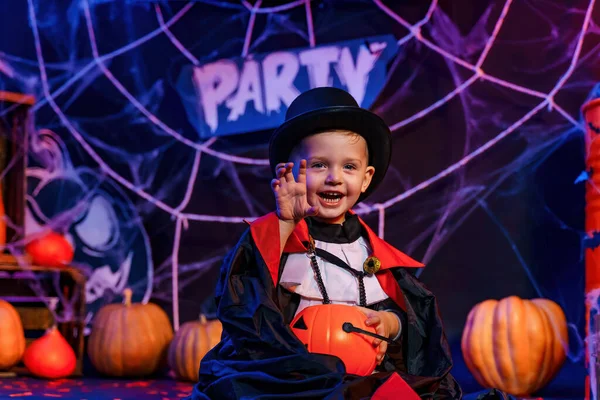 Halloween party. Little cute boy in vampire costume at Halloween party sitting against Halloween decorations and holds pumpkin for candies. Trick or treat