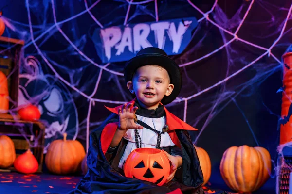 Halloween party. Little cute boy in Dracula costume at Halloween party sitting against Halloween decorations and holds pumpkin for candies.