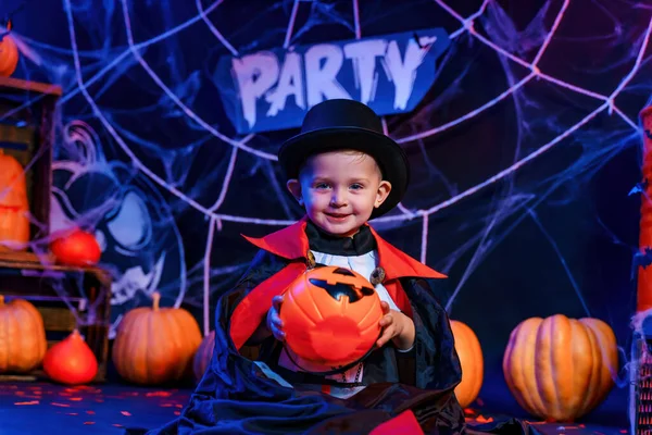 Halloween party. Little cute boy in vampire costume at Halloween party sitting against Halloween decorations and holds pumpkin for candies.