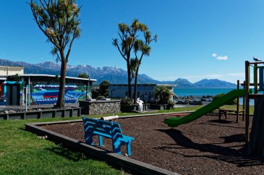 Parlak mavi bir koltuk bir oyun parkında oturur ve tırmanma iskeletinden inen yeşil bir slayta bakar. Tırmanma iskeletinin üzerinde bir seagul var. Oyun alanı Kaikoura 'da denizin hemen yanında, Aotearoa, Yeni Zelanda' da..