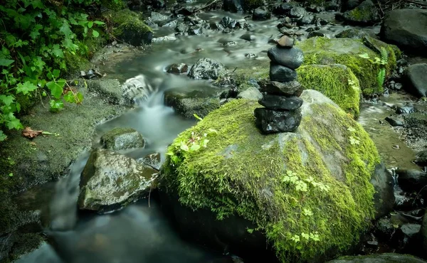 Long exposure of stream with mossy stones and a rock cairn.