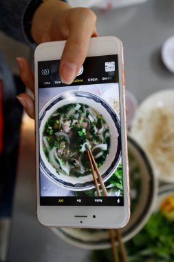 Bir kase Vietnam erişte çorbası, Pho olarak bilinir. Kadın akıllı telefonuyla yemeğinin fotoğrafını çekiyor. Ho Chi Minh Şehri. Vietnam.