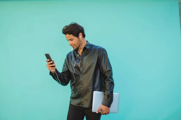 Young male entrepreneur in earphones with netbook watching cellphone during video chat on aquamarine background