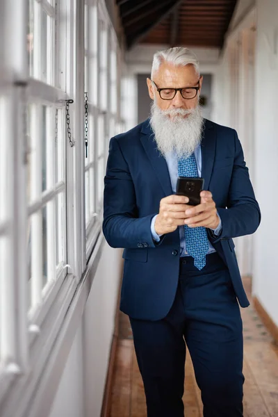 Well dressed male executive with gray beard text messaging on cellphone while standing in passage