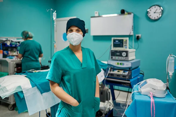 Female doctor in disposable mask standing with hands in pockets while looking at camera against anonymous colleague in hospital