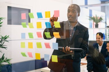 Resmi takım elbiseli, renkli yapışkan notlarla cam duvarın yanında duran ve konferans odasında iş arkadaşlarıyla yaptığı toplantıda bilgi okuyan Afrikalı Amerikalı bir işadamı.