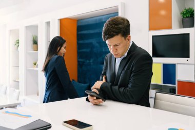 Konsantre iş adamı resmi takım elbiseli, masanın yanında duruyor ve modern ofisteki mesai saatlerinde kadın meslektaşının yanında akıllı telefon kullanıyor.