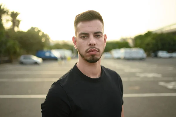 Serious young bearded male in black shirt looking at camera while standing on parking lot on sunny day in city