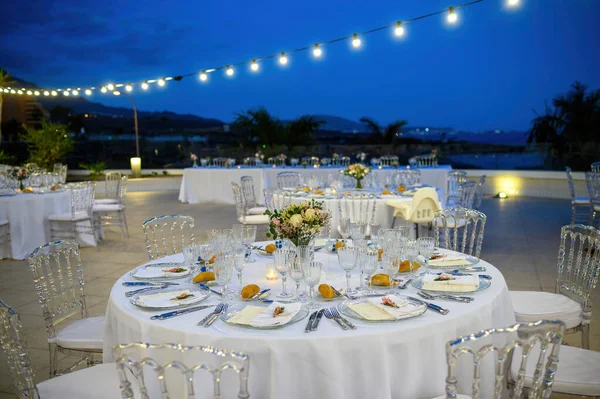 Akşam kutlaması sırasında modern dekore edilmiş açık hava restoranında sandalyelerin yanında cam eşyalar ve beyaz masa örtüsüyle yuvarlak masalar sundu.