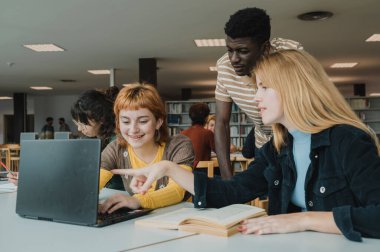Yoğunlaşmış genç kız öğrenci bilgisayar ekranını işaret ederken aynı zamanda çok ırklı sınıf arkadaşlarıyla aynı masada oturuyor ve modern kütüphanede ödevlerini birlikte yapıyorlar.
