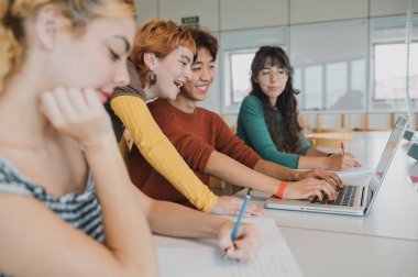 Modern sınıftaki masada otururken günlük giysiler içinde not alan ve dizüstü bilgisayarda yazan pozitif, etnik çeşitliliğe sahip genç grup arkadaşlarının yan bakışı