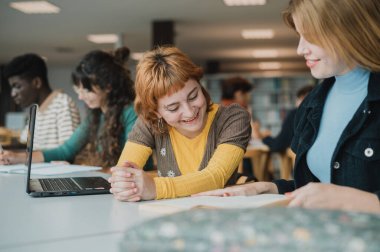 Modern kütüphanede sınav hazırlıkları sırasında masada otururken günlük kıyafetleriyle gülümseyen ve ders kitabı okuyan pozitifler.
