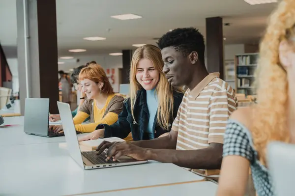 Delighted young multiracial male and female groupmates in casual clothes smiling happily and chatting while typing on laptops during exam preparation in modern light library
