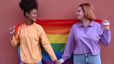 Gerçek zamanlı genç homoseksüel çiftler el ele tutuşup birbirlerine bakarken mutlu bir şekilde duvarın önünde LGBT bayrağıyla dikiliyorlar.