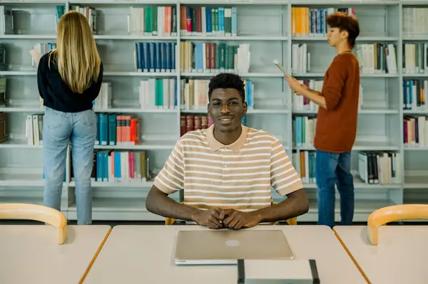Smiling young African American male sitting in library with closed laptop and looking at camera with hands together on desk against bookshelves and fellow students