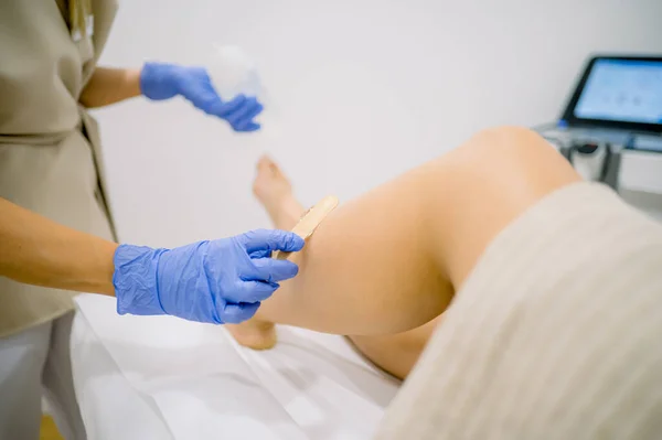 Faceless professional cosmetician in gloves smearing cooling gel on leg of crop client lying on table before IPL procedure