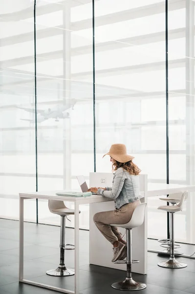 stock image Full body female traveler in casual clothes and straw hat sitting at table and working on remote project on netbook against window with flying plane