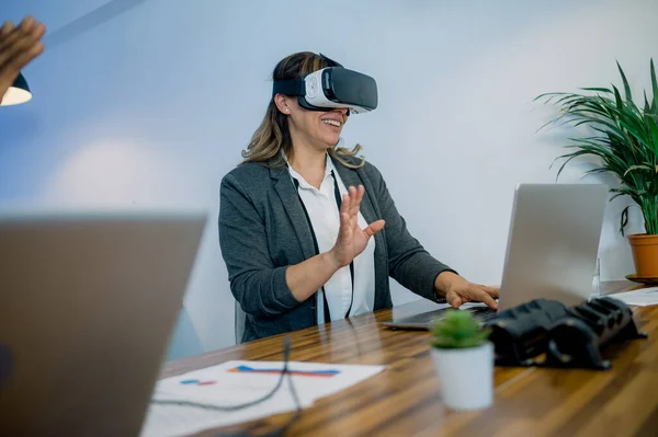 Content middle aged female manager in formal wear smiling and gesturing while experiencing virtual reality in modern headset and using laptop in office