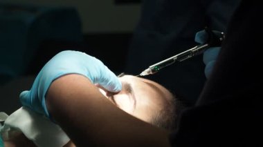 Karanlık ameliyathanede burun estetiği ameliyatından önce kadının alnına ağrı kesici enjekte etmek için lateks eldivenli isimsiz bir doktorun el kamerası görüntüsü.