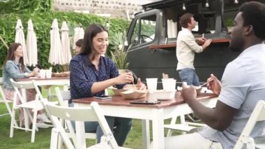 Parkta öğle yemeği sırasında yemek kamyonunun yanında oturmuş salata yerken gülümseyen ve birbiriyle konuşan İspanyol bir adamın el kamerası görüntüsü.
