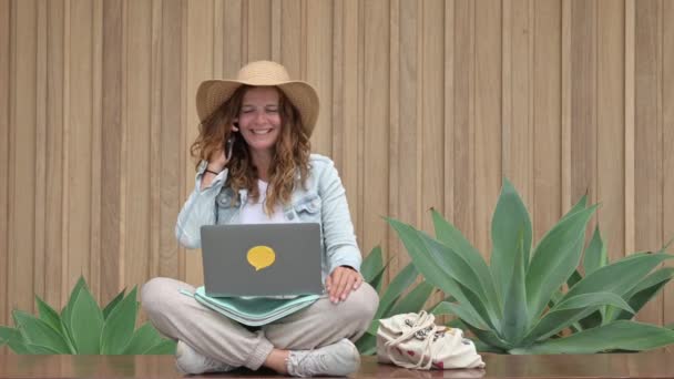 携帯電話で話すサンハットを持つカジュアルな服の幸せな女性は リゾートでのリモートプロジェクトの作業中にエキゾチックな植物の近くの国境に足を踏んで座っている間 その後 ネットブックを使用し続けます — ストック動画