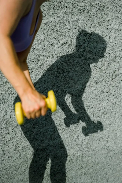 Güneş ışığında gri beton duvara karşı egzersiz yapan yüzü olmayan, spor ayakkabılı bir kadının gölgesi.