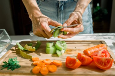 Tahta kesme tahtasıyla sulu kiviyi soyarken mutfakta sağlıklı salata için malzemeler hazırlayan anonim bir kadın.