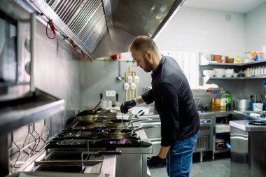 Ciddi sakallı, sıradan kıyafetler giymiş ve eldivenli bir aşçının parlak, modern bir restoranın mutfağında tavada lezzetli yemekler kızartışının yan görüntüsü.