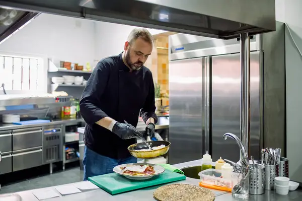 Sakallı, odaklanmış, günlük giysiler içinde, eldivenli, modern bir restoranda tavadan tabağa lezzetli yemekler servis eden erkek şef.