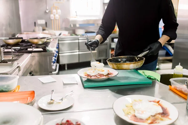 Crop male chef in black uniform with pan serving hot fried egg on bacon for breakfast in modern kitchen of restaurant