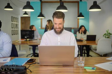 Ciddi sakallı adam dizüstü bilgisayarla masada oturuyor ve modern çalışma alanında çalışırken kameraya bakıyor.