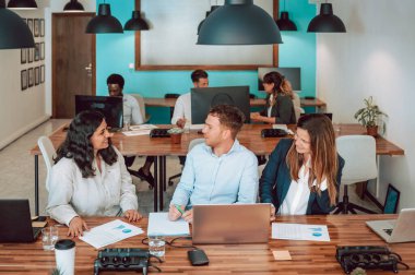 Bir grup iş arkadaşı masada dizüstü bilgisayarlarla ve belgelerle oturuyor ve açık modern ofiste proje üzerinde çalışırken birbirlerine bakıyorlar.