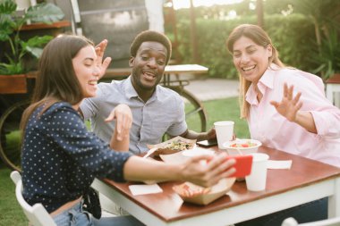 Mutlu erkek ve kadınlar gündelik giysiler içinde el sallıyor ve masada otururken gülümsüyor ve sokak restoranında yemek sırasında video görüşmesi yapıyor.