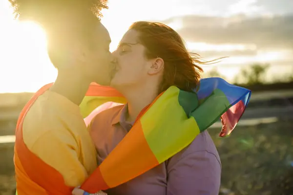 Side view of woman and woman with rainbow flag hugging and kissing each other while standing on blurred background