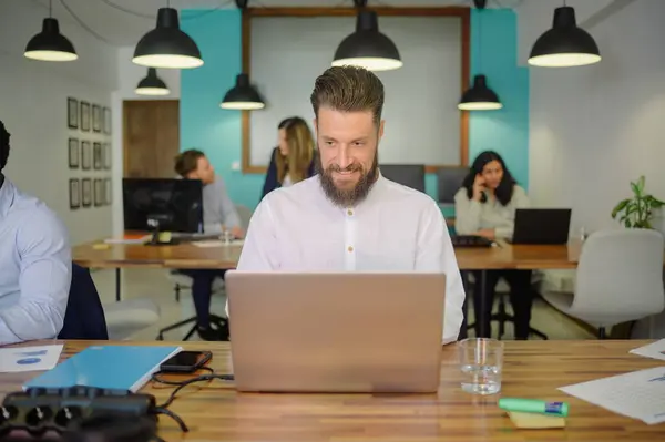 Serious bearded man sitting at table with laptop and looking at camera while working in modern workspace