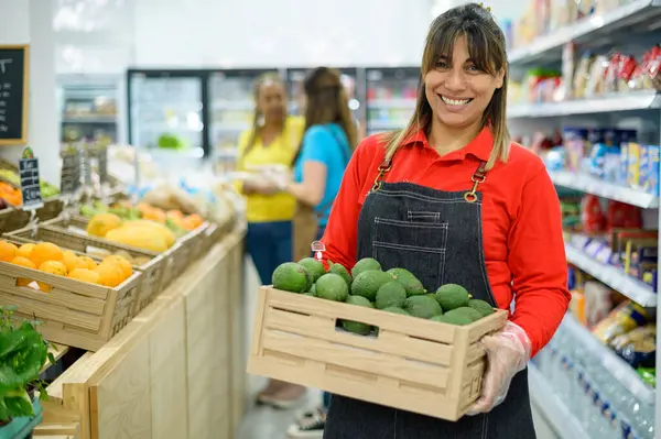 Positive adult Hispanic saleswoman in apron and plastic gloves carrying box with fresh avocado while working in grocery store with shelves of various goods and looking at camera