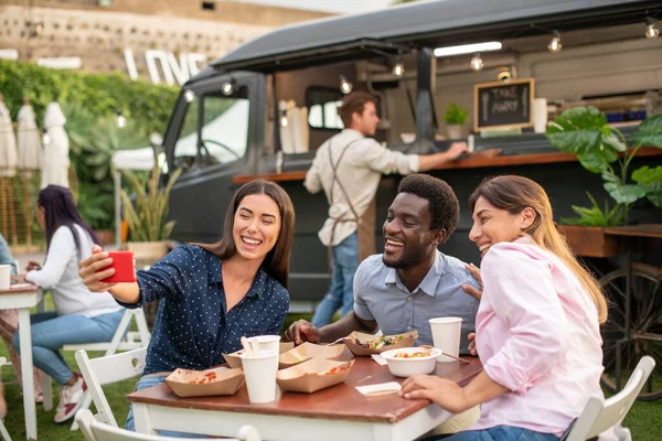 Group of happy multiracial friends gathering at table with delicious takeaway meal and taking self portrait against food truck in park