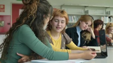 Laptop 'la masada oturup kütüphanede ödevlerini yaparken günlük kıyafetleriyle gülümseyen ve birbiriyle konuşan pozitif, etnik çeşitliliğe sahip genç bayan arkadaşlar.