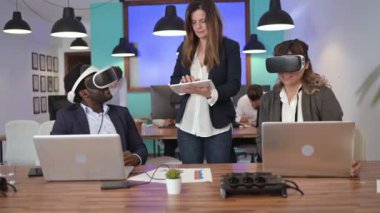 VR gözlüklü çok ırklı iş arkadaşlarını, modern ofiste netbook 'larla sanal gerçekliği keşfederken gerçek zamanlı bir kadın tableti karıştırıyor.
