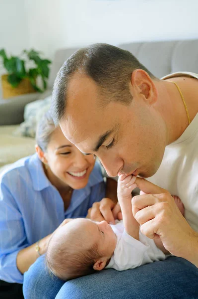 Happy father in beige t shirt kissing arms of adorable newborn baby near smiling wife in living room