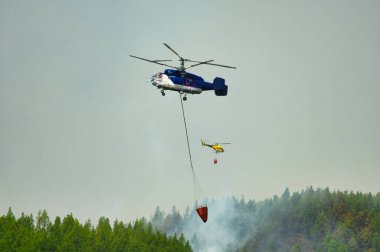 Yeşil ormanın üzerinde uçan helikopter ve Tenerife 'de yaz günü bulutlu gökyüzünün altında söndüren yangın