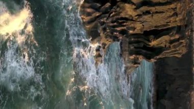 Dalgalı köpüklü dalgalar Tenerife kırsalındaki bulanık arka planda gün ışığında dağdaki kayaların keskin kenarlarına çarpıyor ve deniz suyunu kırıyor.