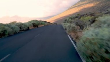 Boş asfalt yolda gerçek zamanlı hareket. Tenerife 'nin yol kenarında yeşil çalılar ve işaretler var. Güneşli gün ışığında dağların tepesinde beyaz bulutlara karşı.
